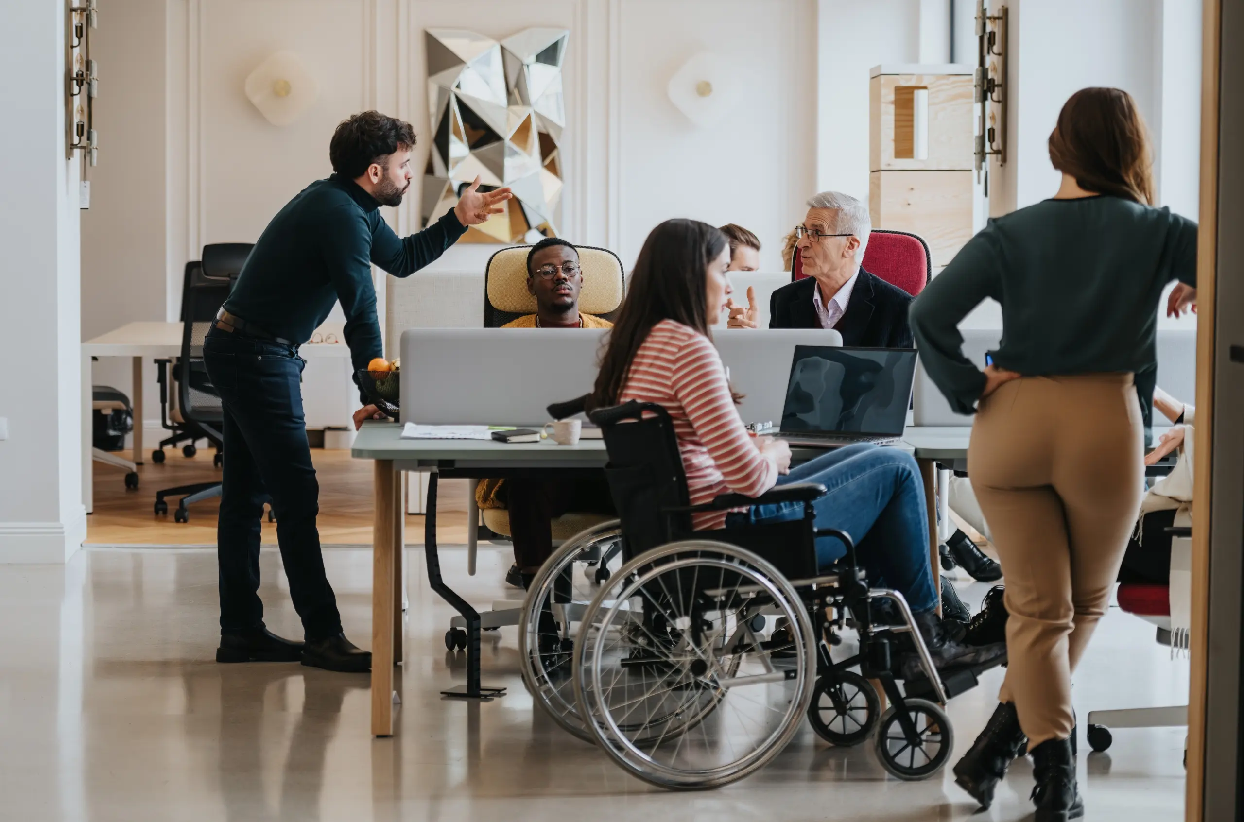 Office with several people with one standing man talking to two seated people, there is one lady on a wheelchair and another lady standing up