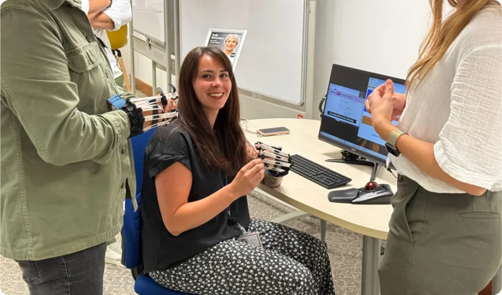 Female presenter demonstrating an empathy workstation to two employees, one seated and smiling to the camera and one standing, both employees are wearing mobility simulation gloves