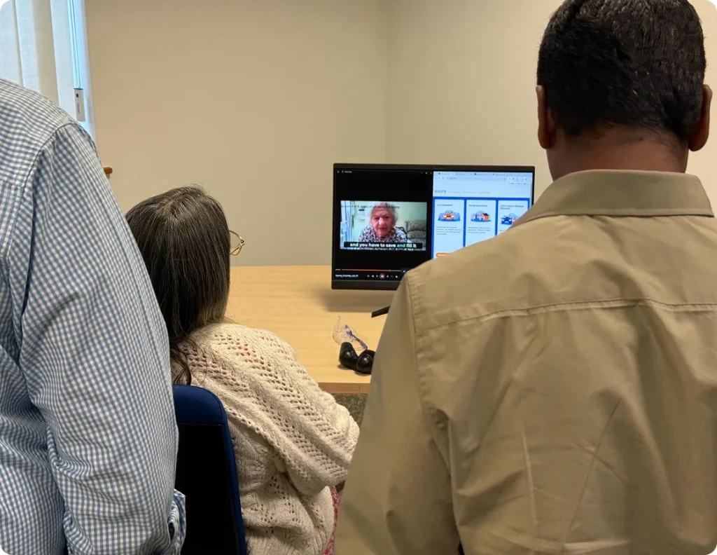 Three people watching a video of an elderly woman at an ageing empathy workstation