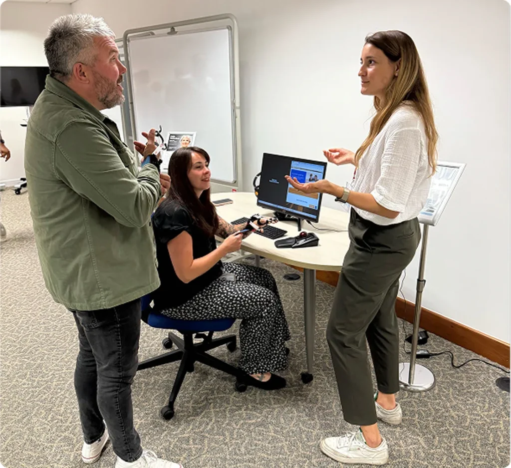 Female presenter demonstrating an empathy workstation to two employees, one seated and one standing, both employees are wearing mobility simulation gloves