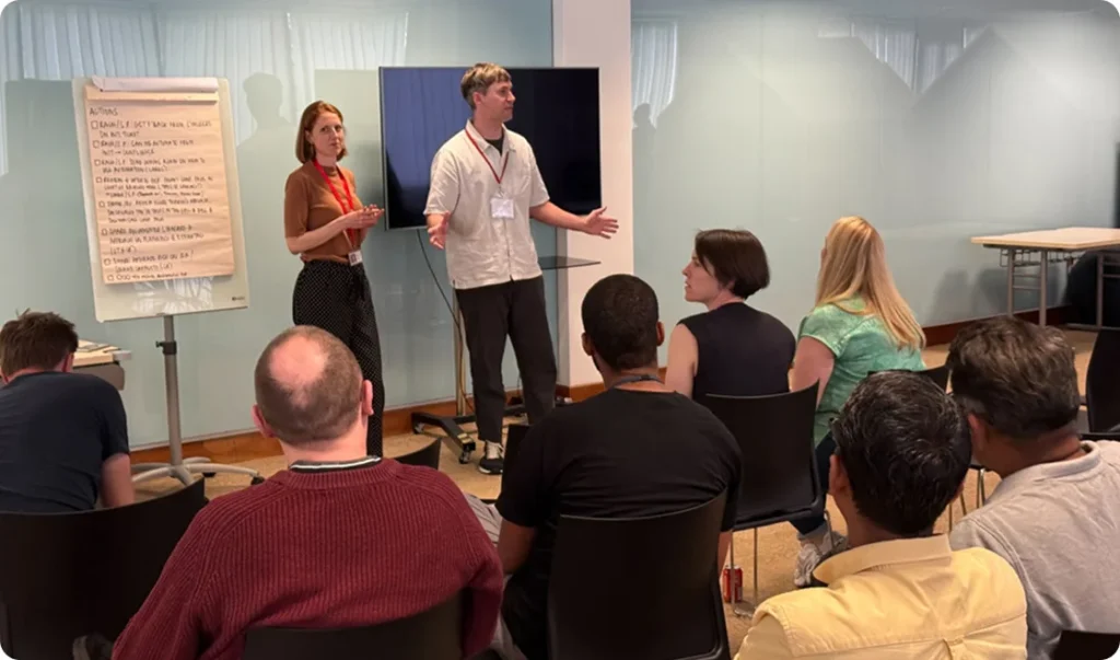 Male and female presenters standing before a seated audience, with a large monitor and flipchart