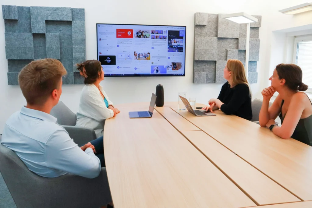 Four people seated by a large conference table with two laptops facing a large monitor showing a webpage.