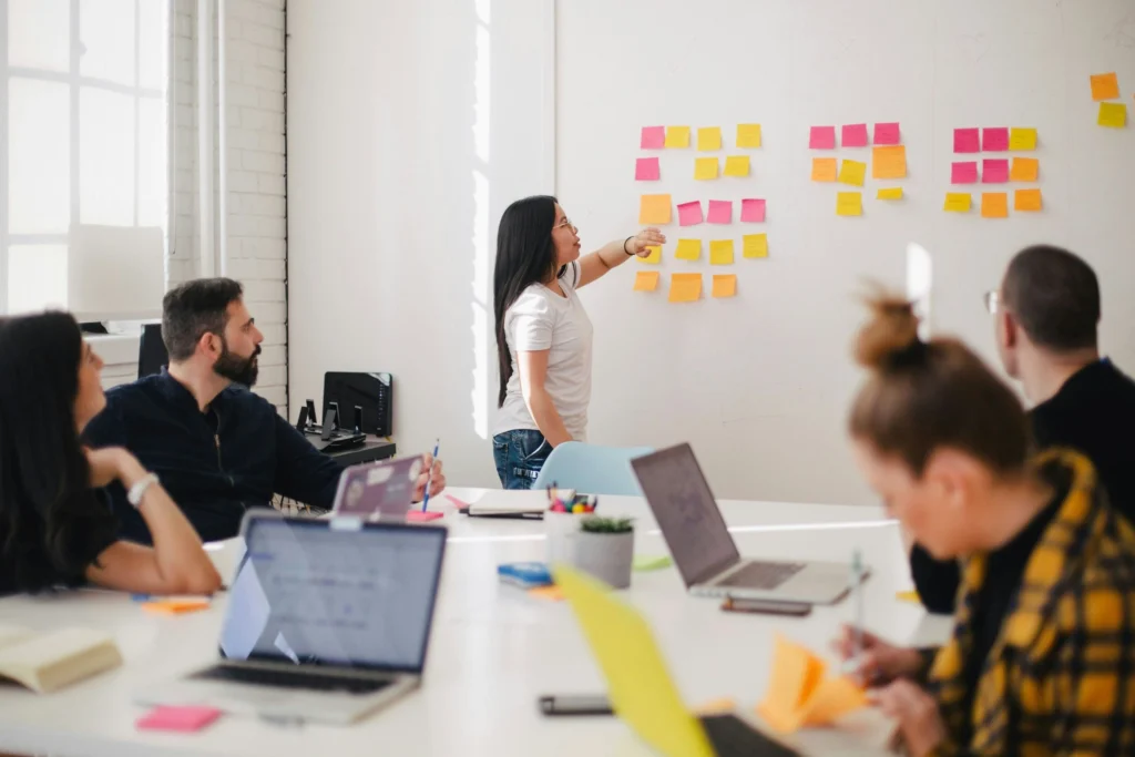 Meeting room with large table with young professionals on their laptops watching a young lady point to a wall of post-it notes
