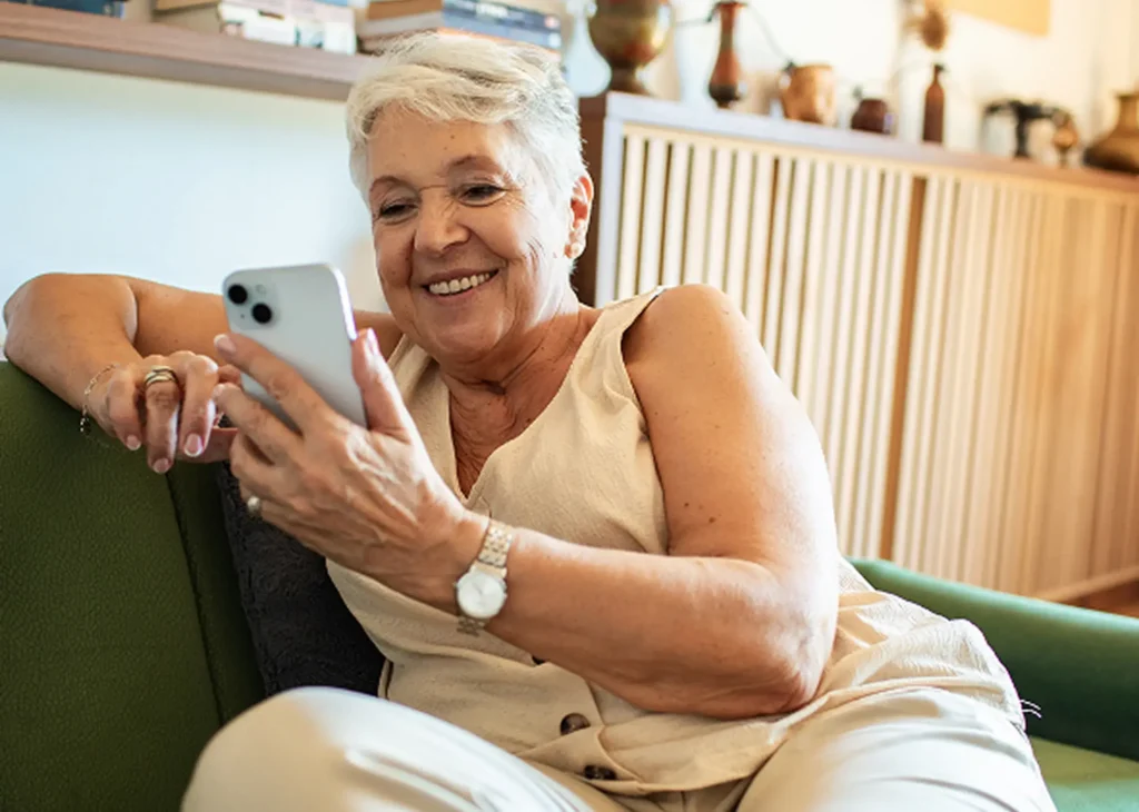 Elderly lady seated sideways on a green sofa and smiling at a smartphone she is holding