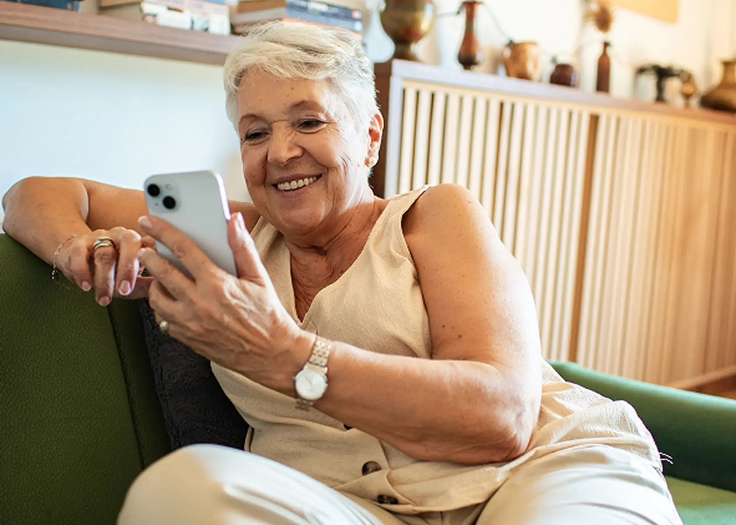 Elderly lady seated sideways on a green sofa and smiling at a smartphone she is holding
