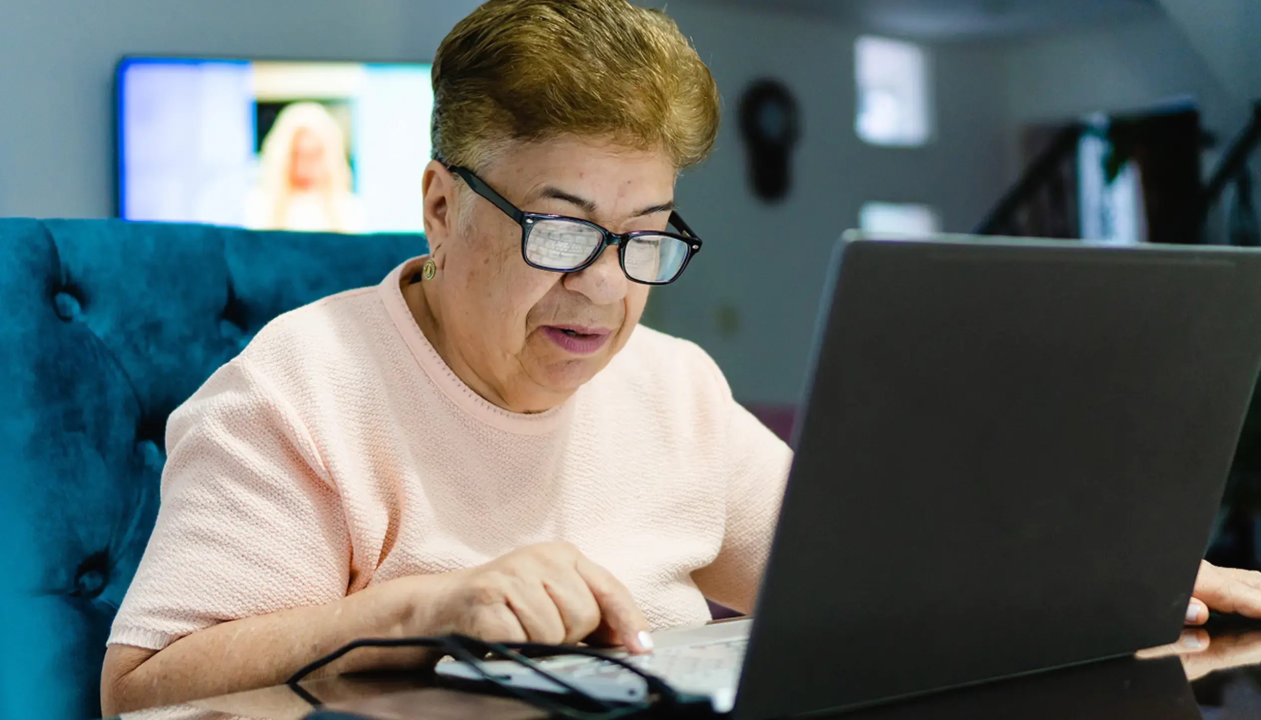 Older woman wearing glasses using a laptop at home, concentrating on the screen with assistive technology nearby.