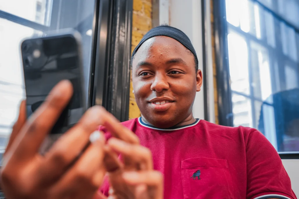 A person smiling while using a smartphone, seated near a window in a casual setting.