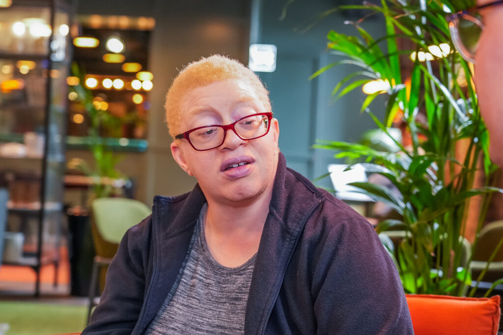 A person wearing red glasses and a dark jacket is talking to someone in a brightly lit indoor setting with green plants and blurred lights in the background.