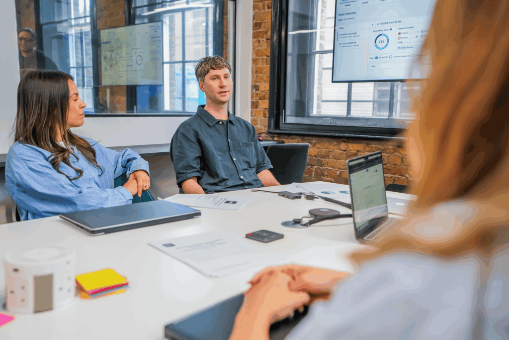 A group of colleagues in a meeting room discussing accessibility metrics displayed on a screen.