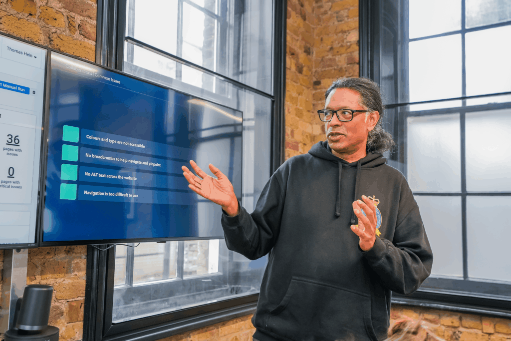 A man in a black hoodie gives a presentation on digital accessibility issues, standing beside a screen displaying common accessibility problems.