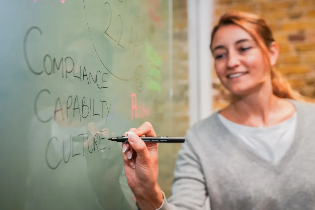 A women writing the words "COMPLIANCE, CAPABILITY, CULTURE" on a transparent board with a black marker.