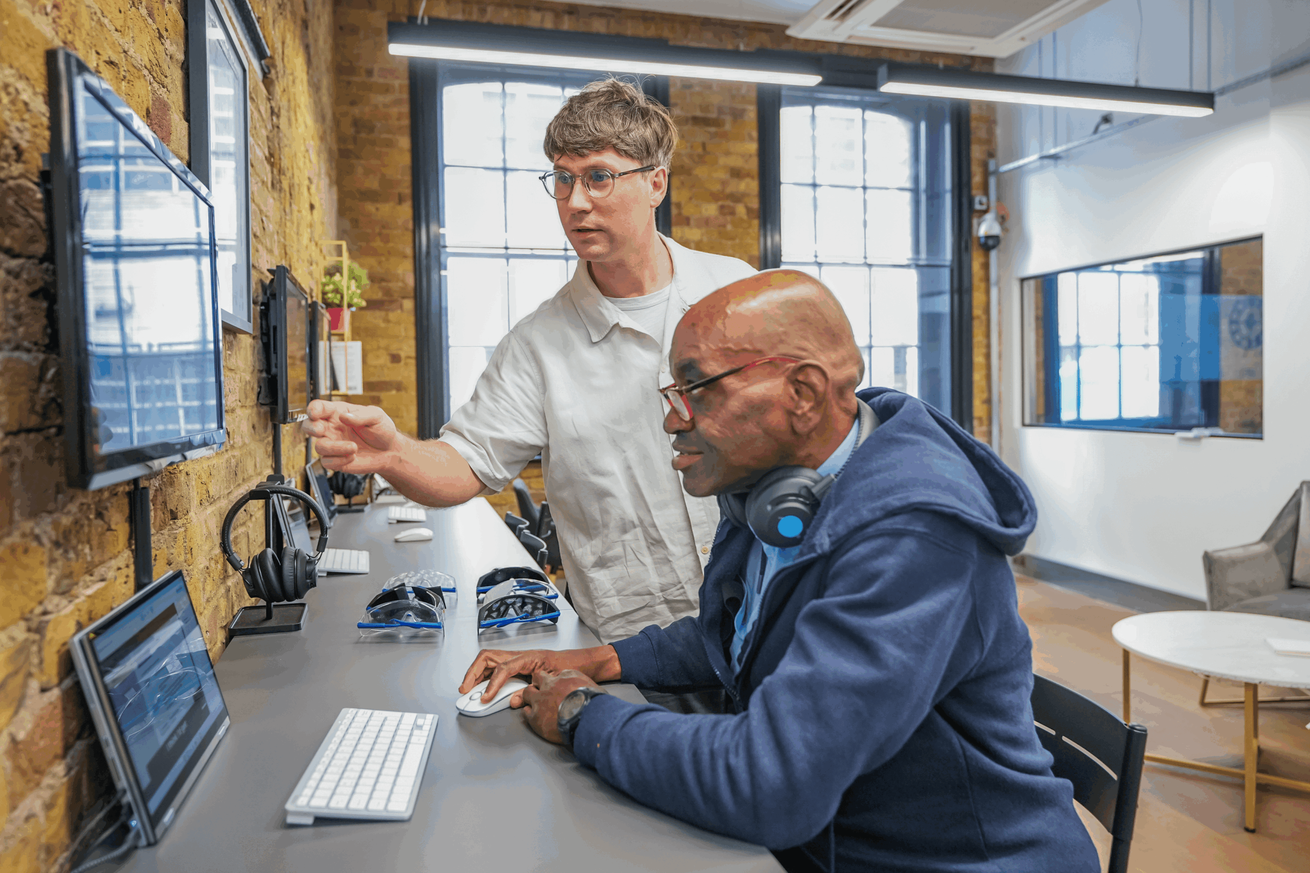 Two people collaborate at a computer workstation, with one person guiding the other by pointing at the screen.