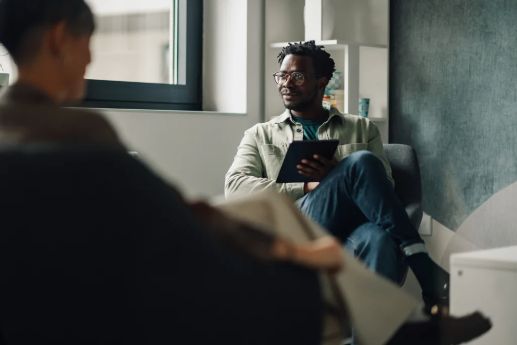 Man sitting in a chair holding a tablet, attentively listening during a one-on-one conversation in a modern office setting.