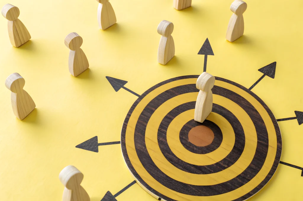 Wooden figure standing at the centre of a yellow and black target, surrounded by arrows pointing outward and other wooden figures on a yellow background.
