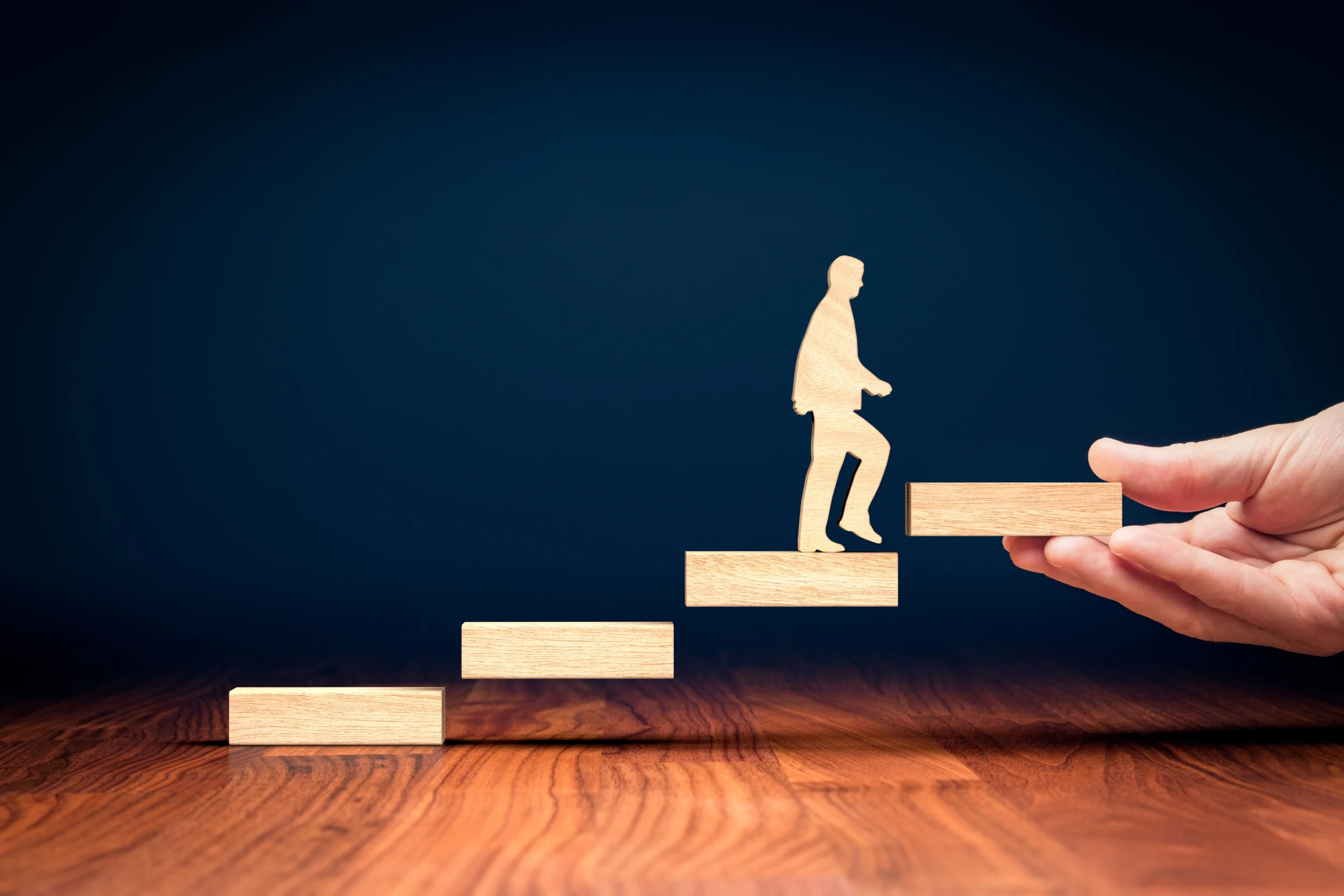 A wooden figure walking up ascending wooden steps, with a hand supporting the final step, symbolising support and growth.