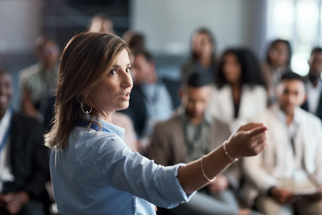 A woman speaking confidently to a diverse audience during a leadership session or corporate event.