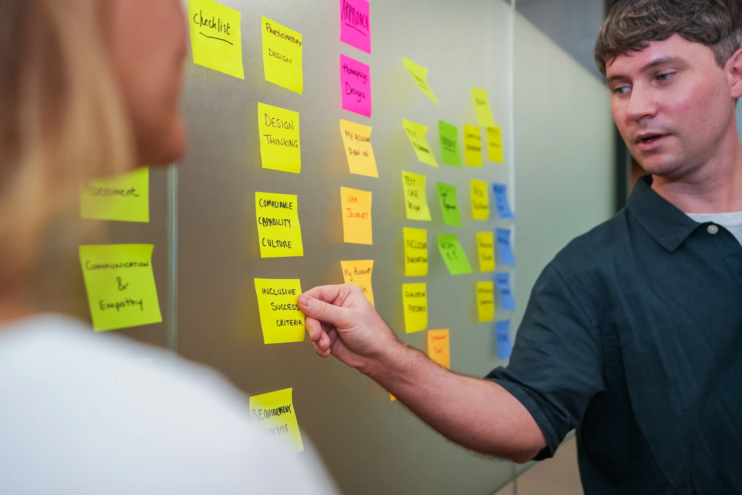 A man leads a planning session, pointing at a wall covered in brightly coloured sticky notes with terms like "compliance," "empathy," and "inclusive success criteria."