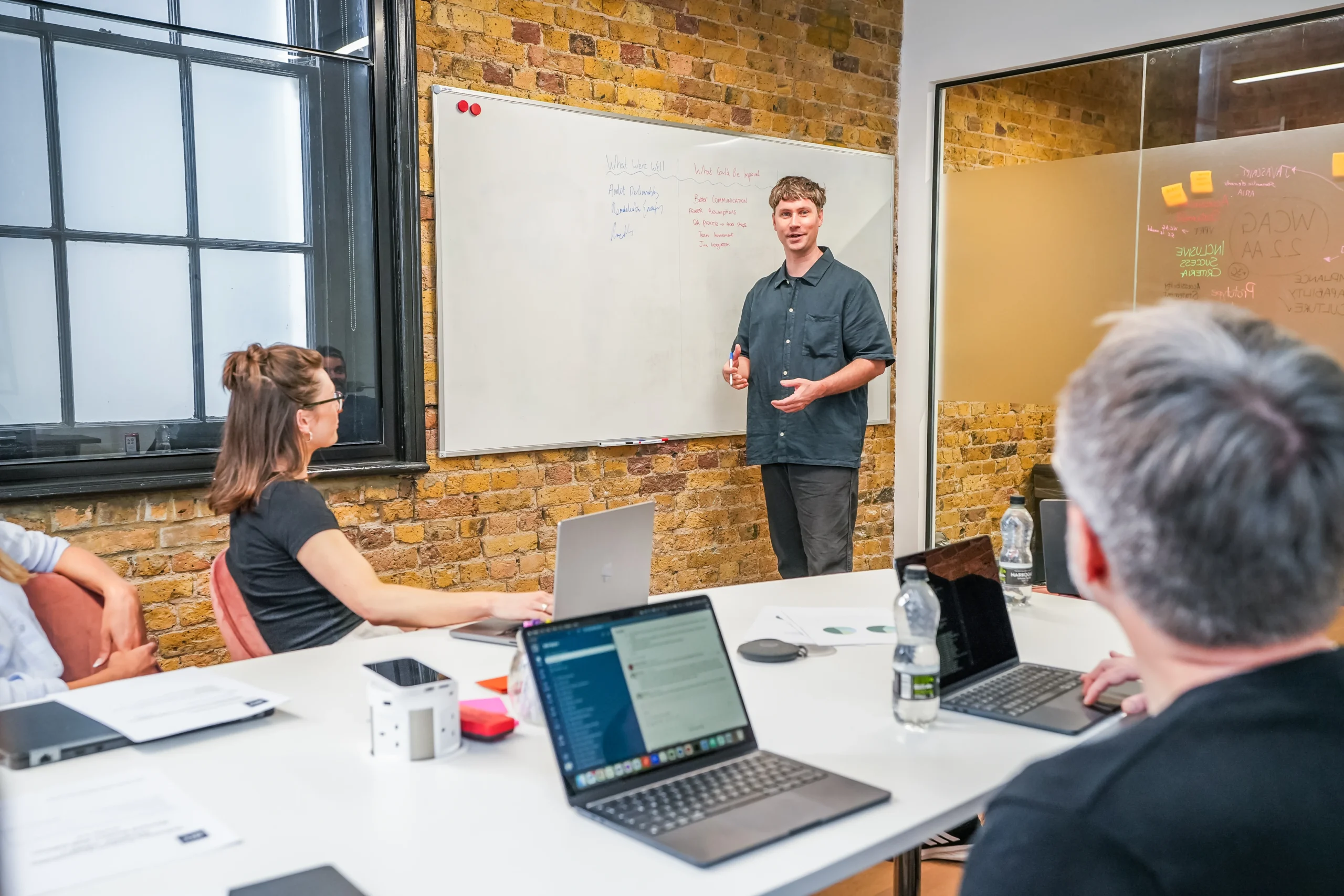A man delivers a presentation at a whiteboard in a modern meeting room while colleagues seated at a table listen and take notes on laptops.