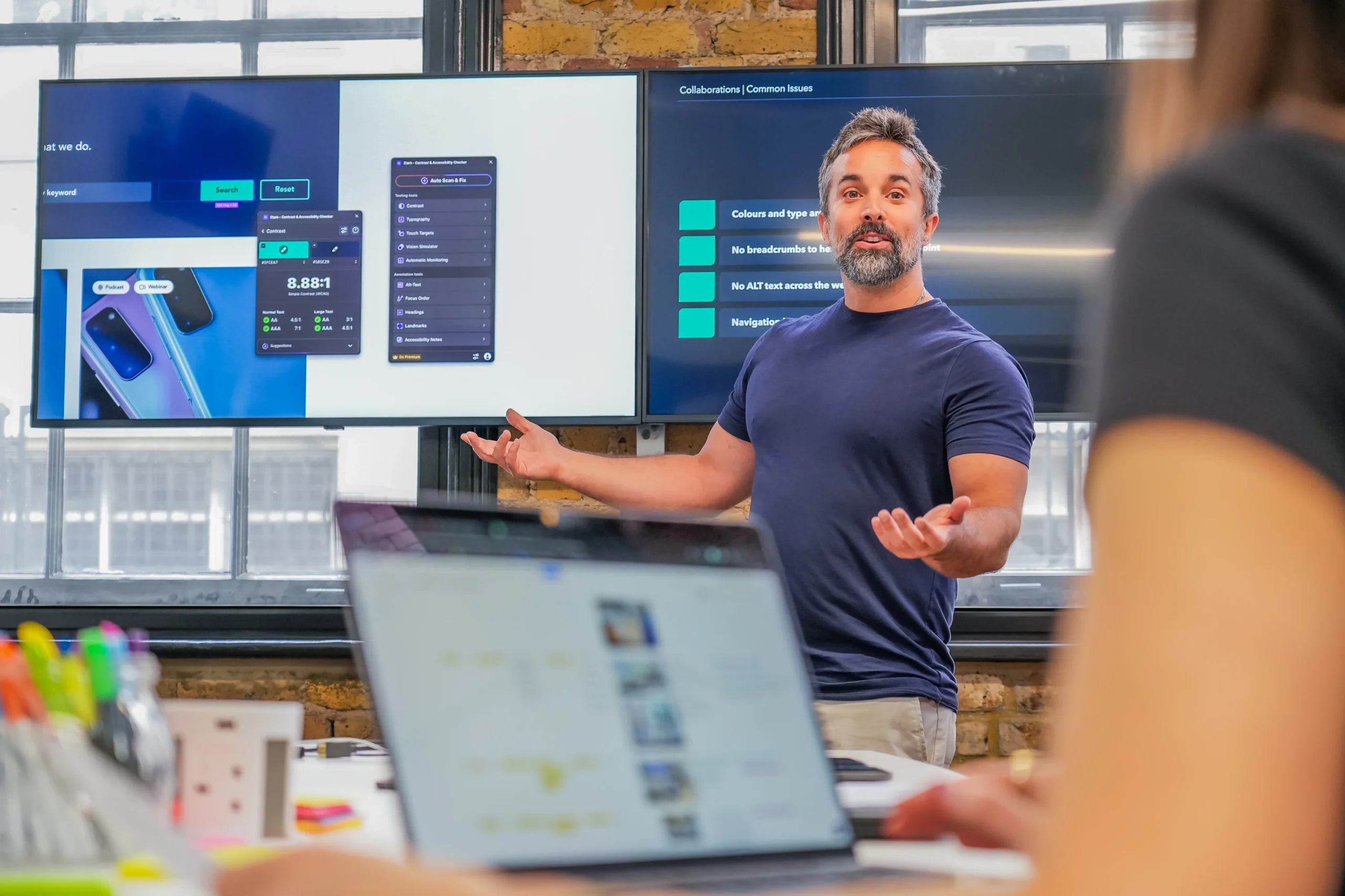 Presenter delivering an accessibility consulting session in front of large screens showing a web interface and audit results within Figma.