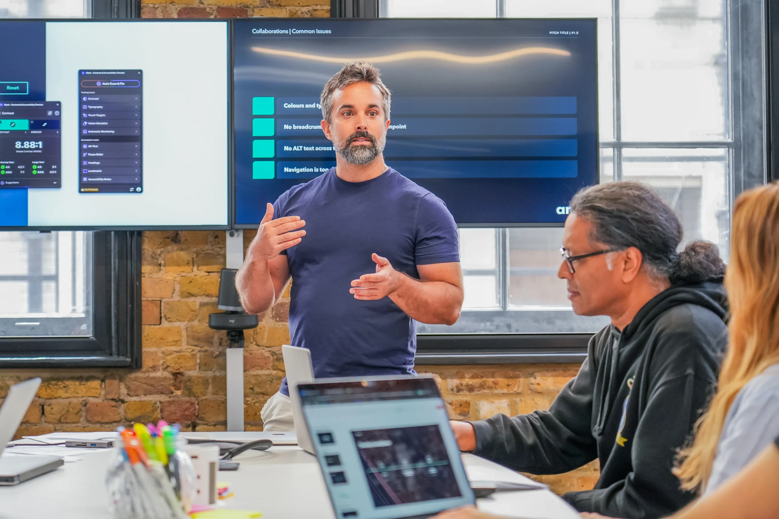 A designer leads an accessibility training session, gesturing as team members listen, with example UI issues displayed on a screen behind him.