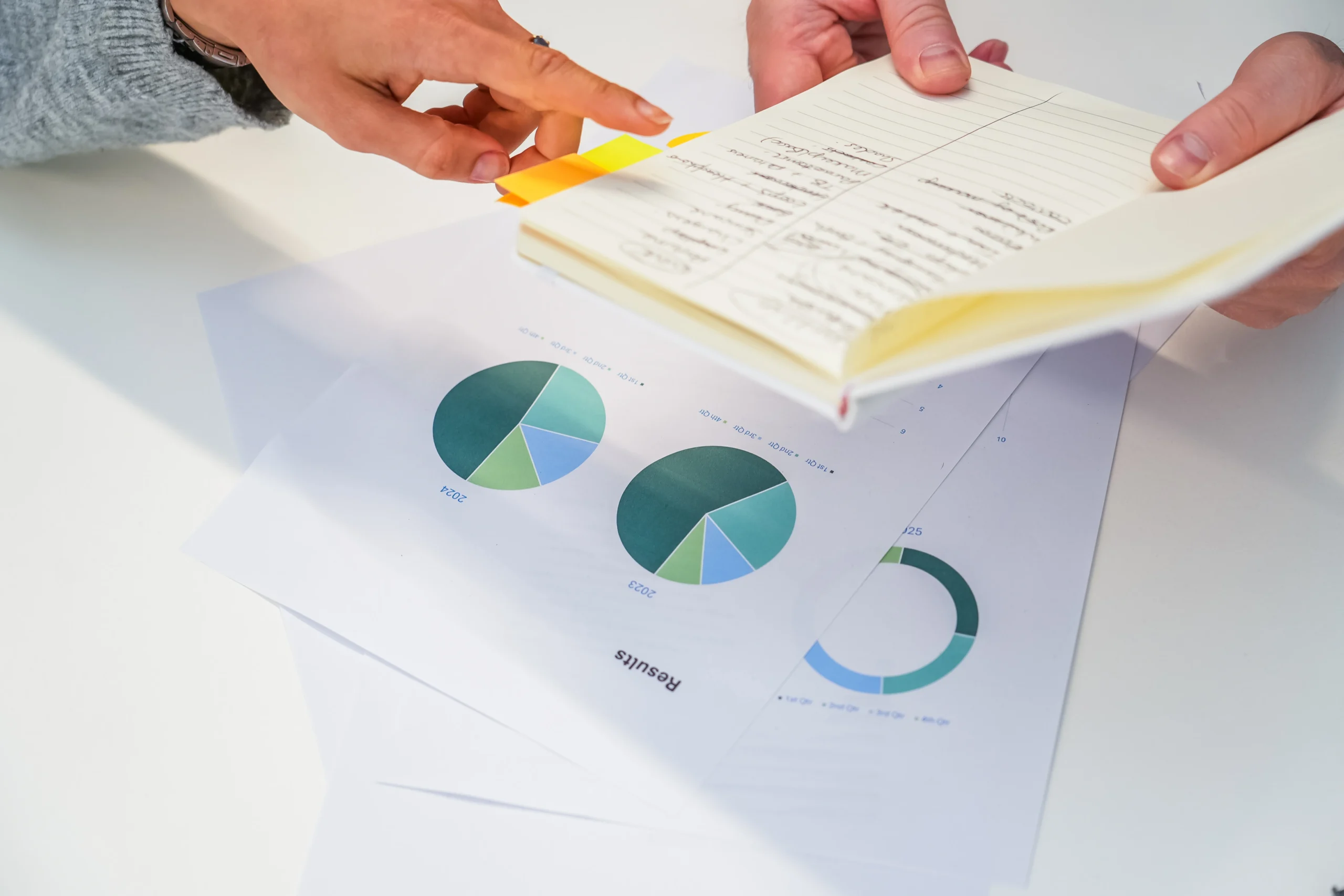Close-up of hands pointing to notes in a notebook and reviewing pie chart reports on a desk, showing data analysis in progress.