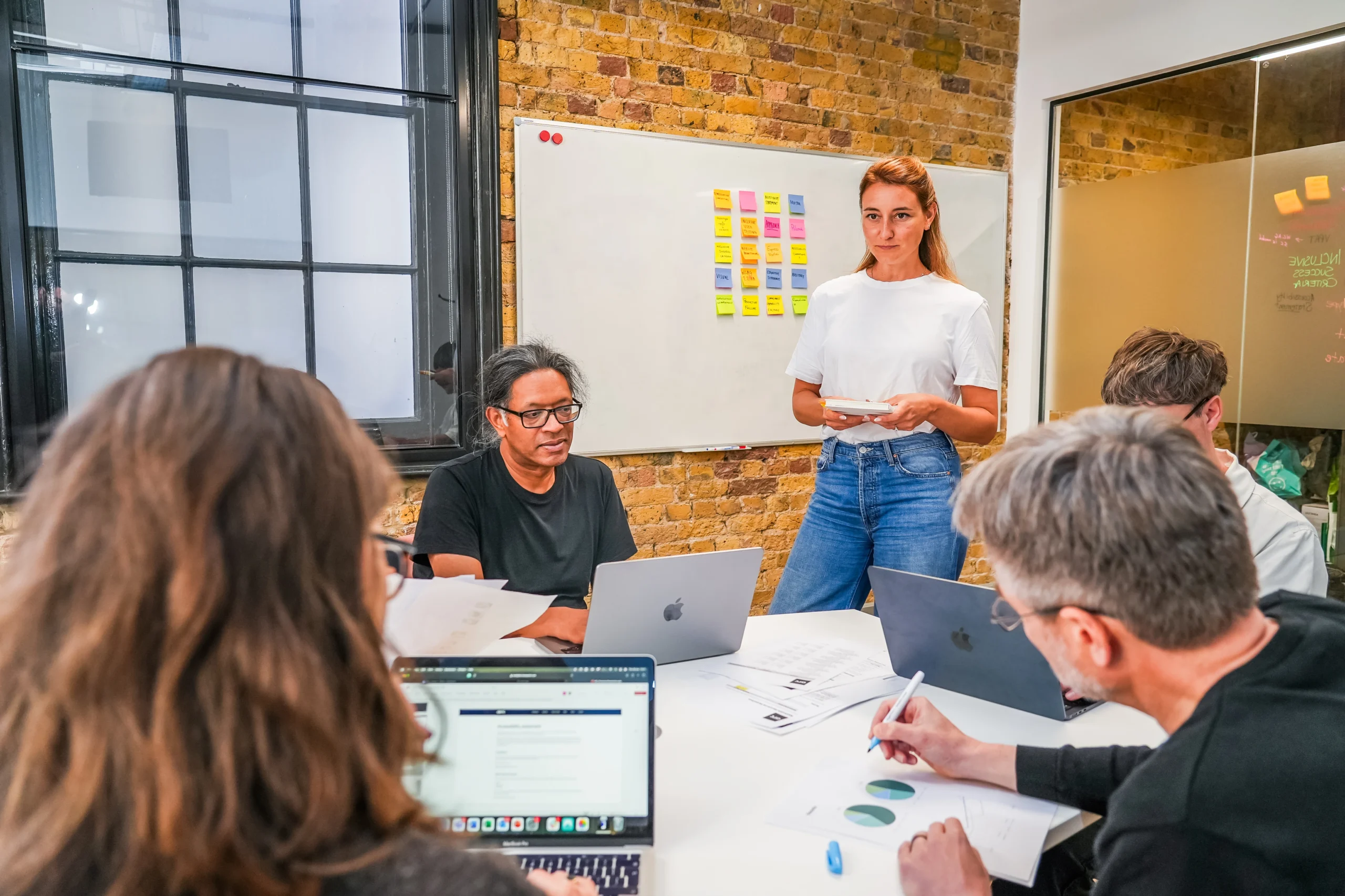 A group of people sit and stand around a table in a modern office, reviewing charts and sticky notes as part of a collaborative workshop.