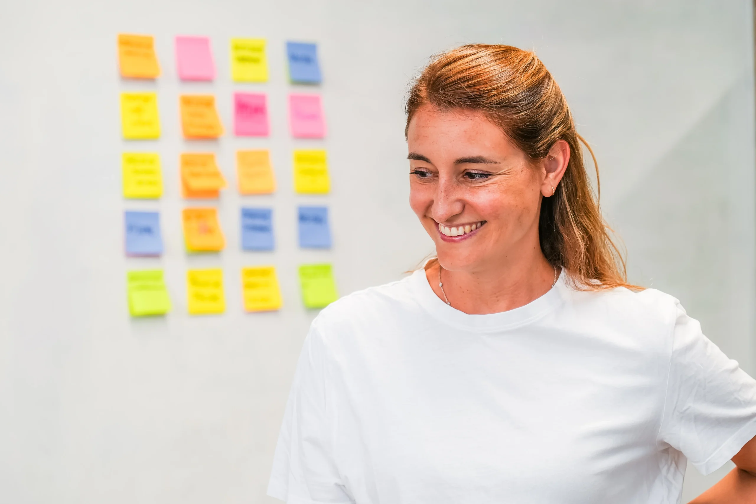 A woman in a white shirt smiles while standing in front of a wall covered in colourful sticky notes arranged in a grid.