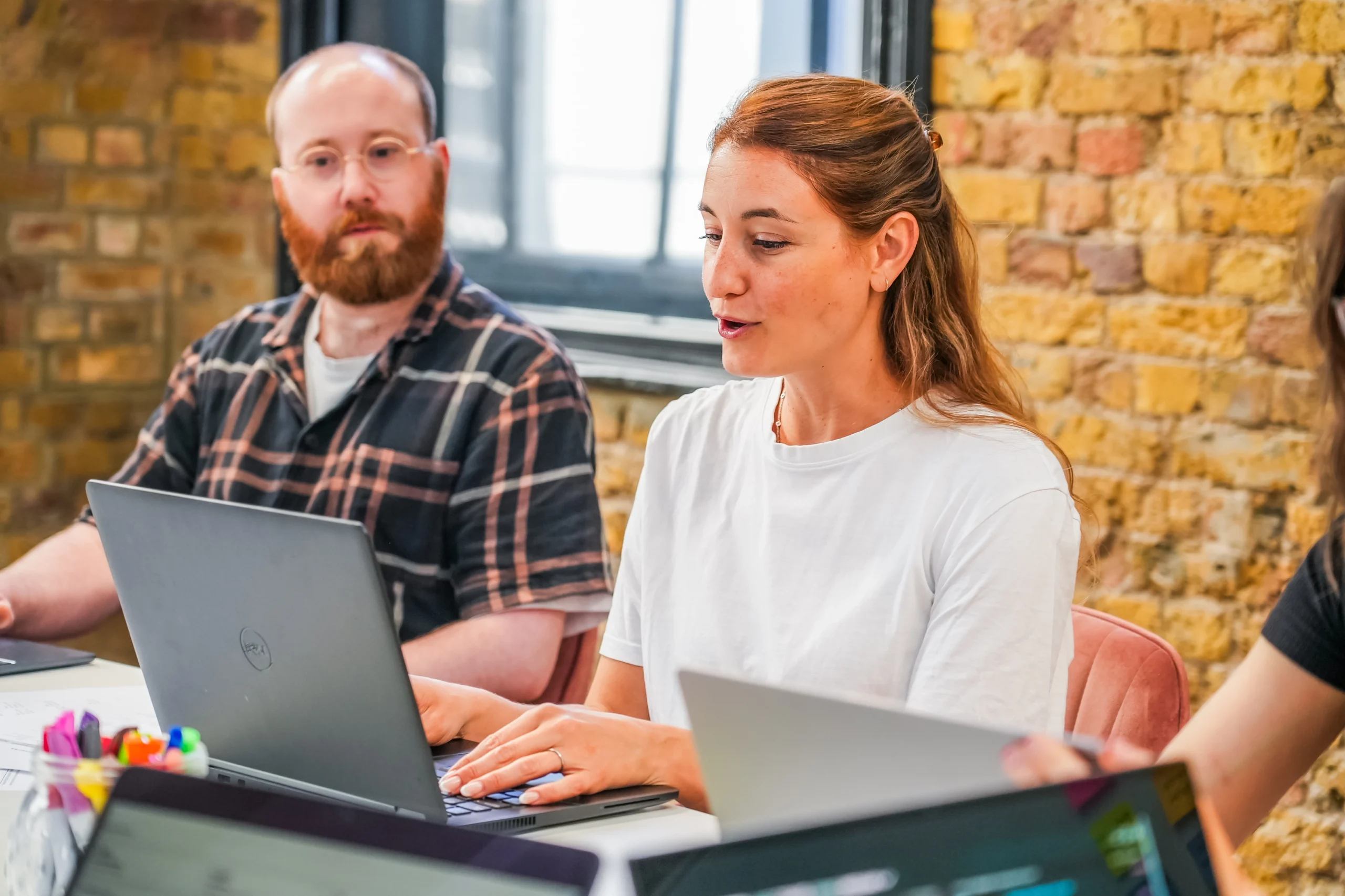 Two team members at laptops in a meeting room discussing digital remediation strategies, one speaking while others listen.