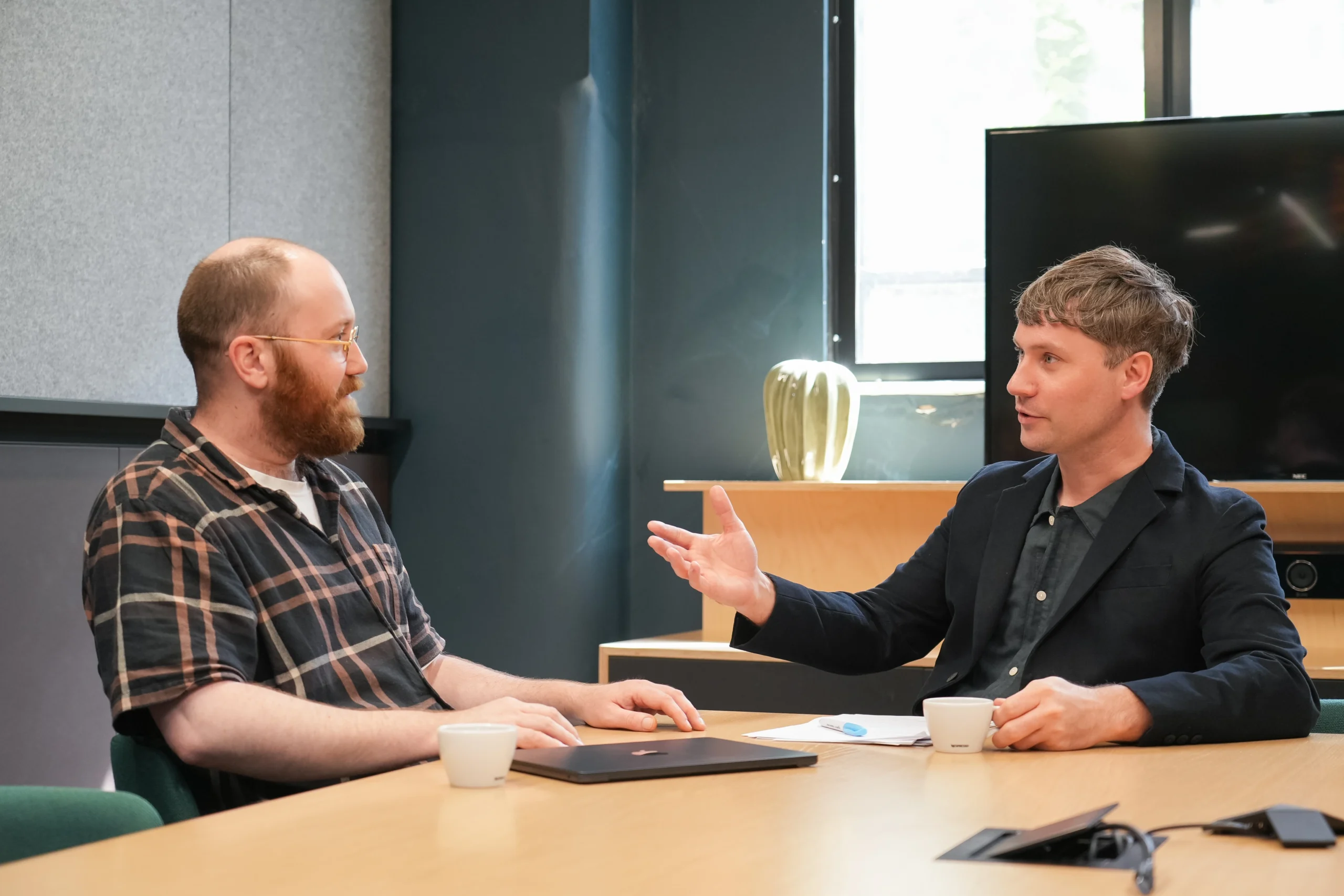 Two men sit across from each other at a conference table in discussion, one gesturing as they talk. They have laptops and coffee cups in front of them.