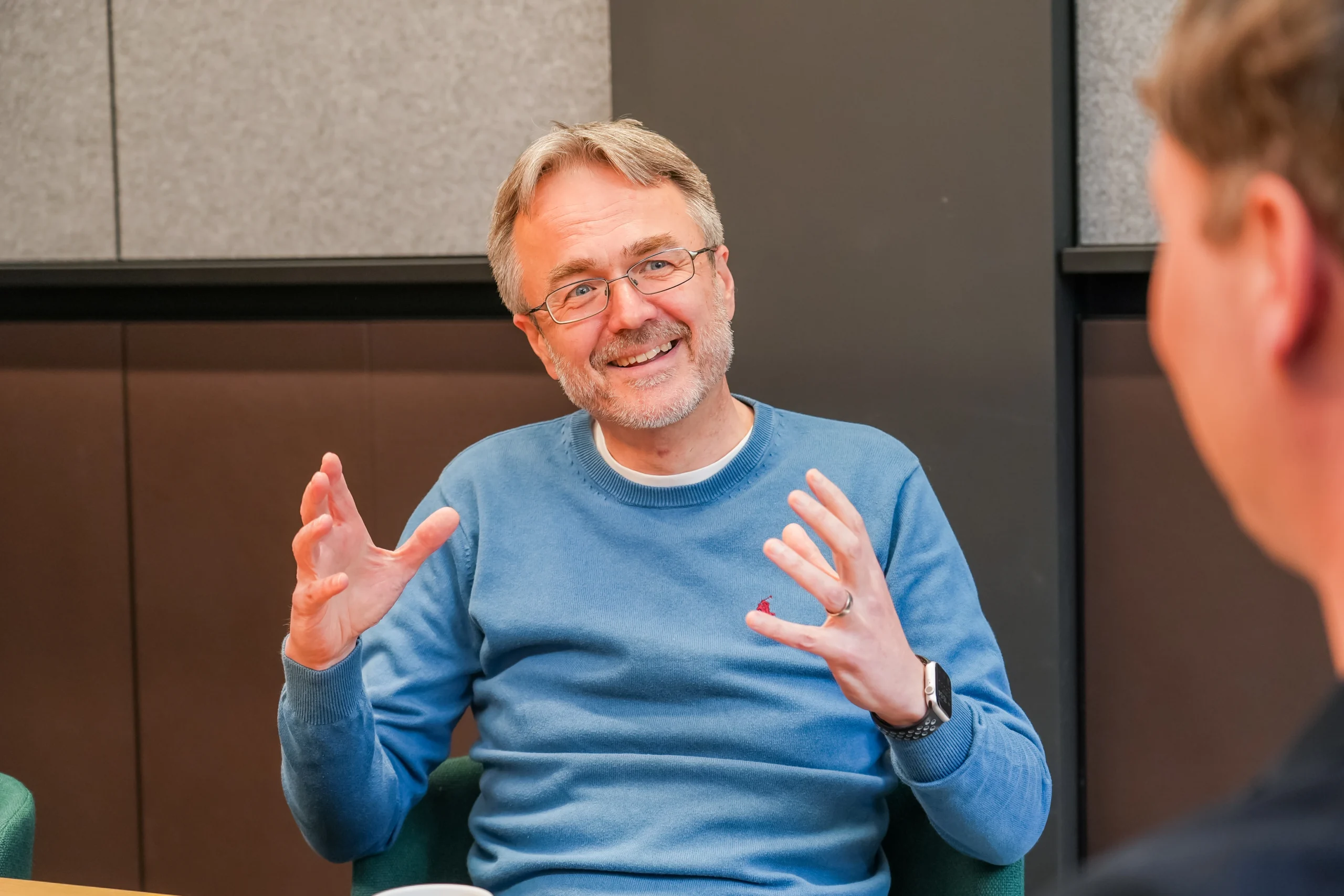 An older man wearing a blue jumper gestures expressively while smiling during a conversation, seated at a table in a modern office setting.