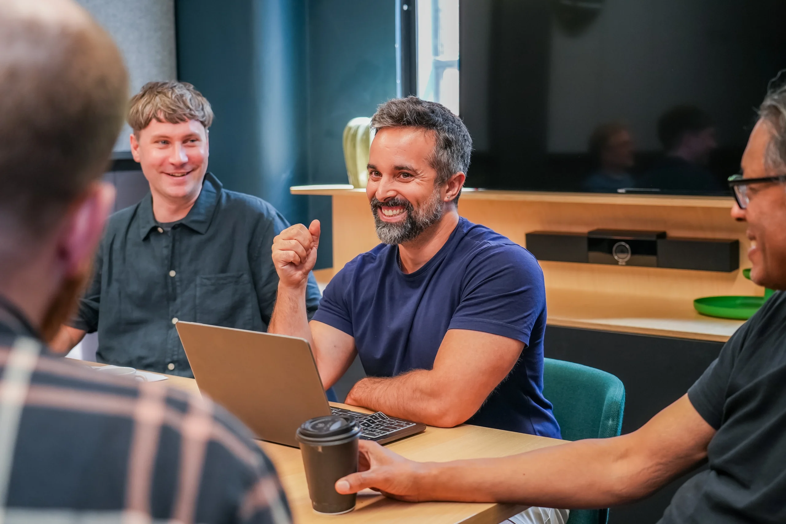 A group of professionals sit around a table in a meeting room, smiling and engaged in conversation. One man gestures confidently while speaking, with a laptop in front of him.