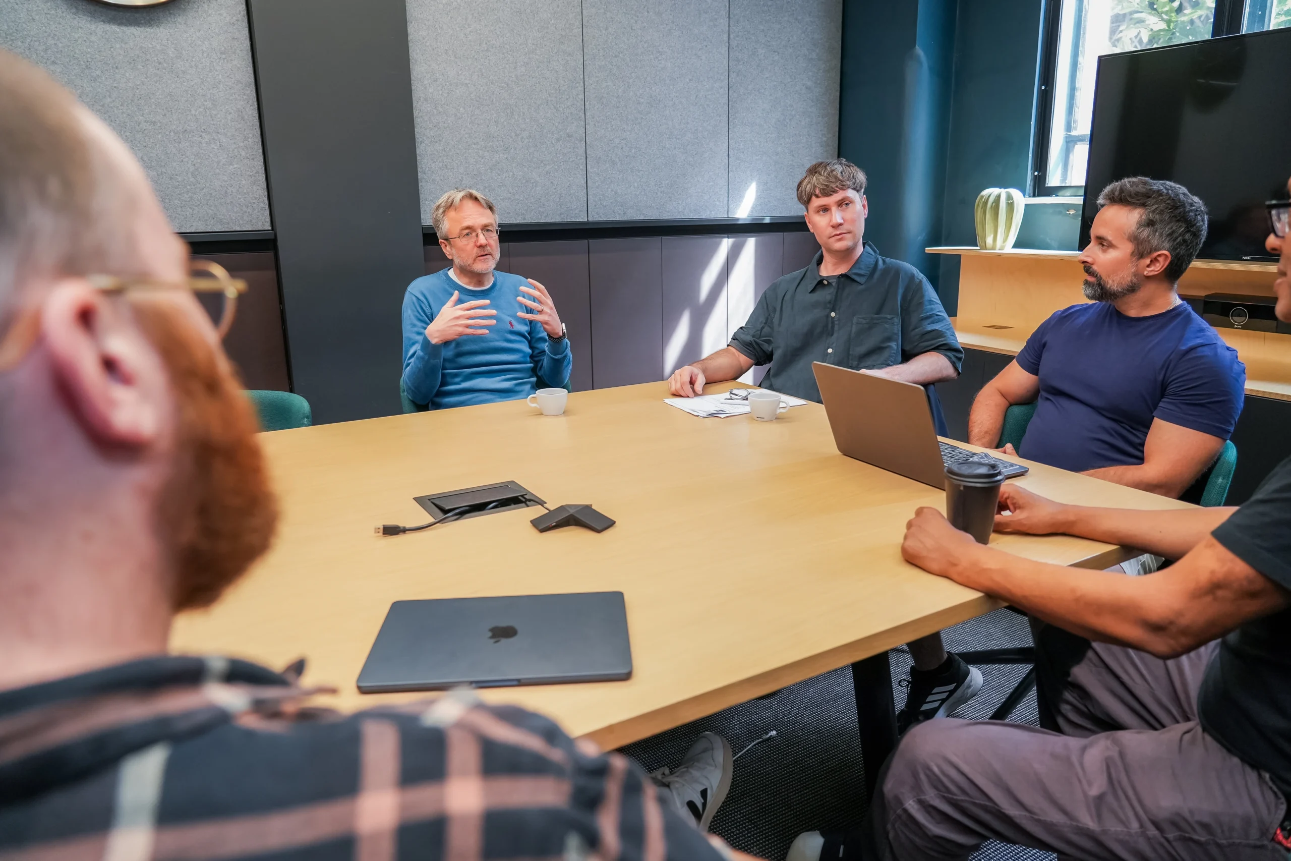 A team of five professionals having a focused discussion around a boardroom table with laptops, notebooks, and coffee.