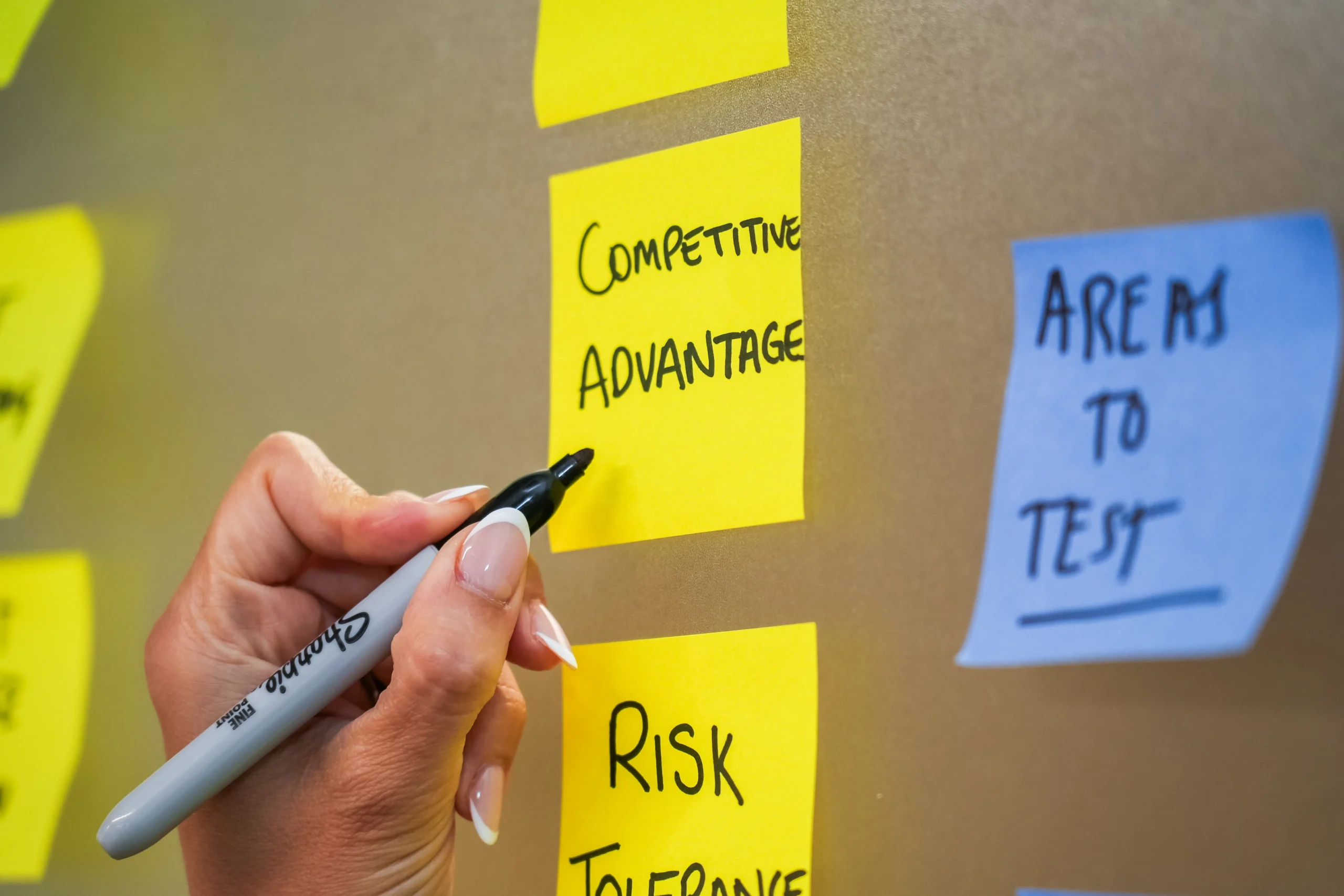 A hand writes “Competitive Advantage” on a yellow sticky note with a black Sharpie.