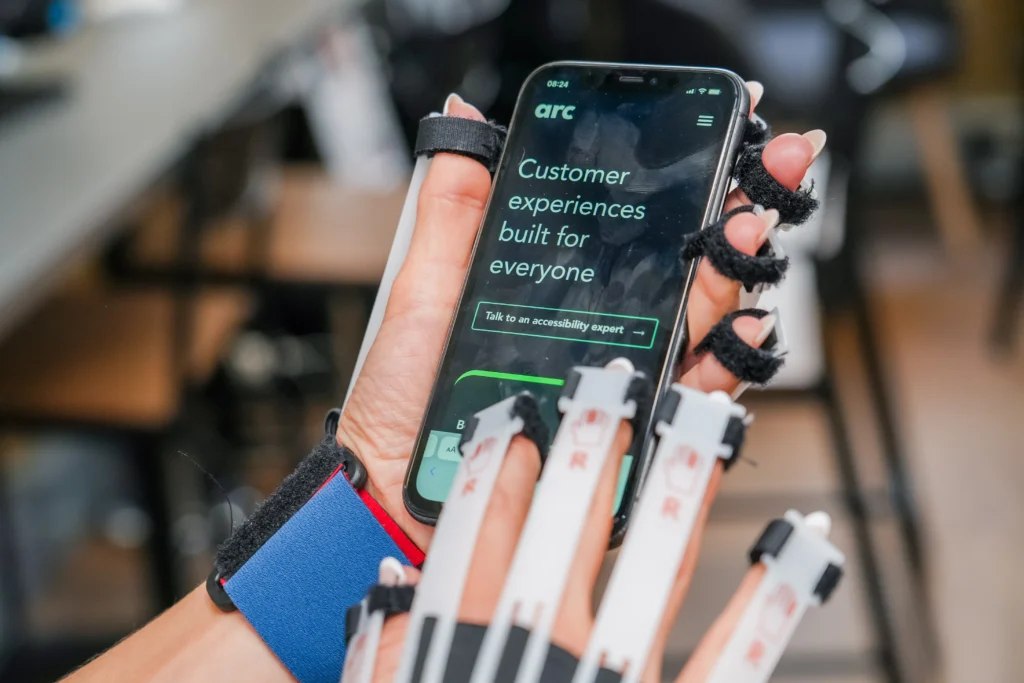 Close-up of a person wearing Cambridge simulation gloves interacting with a smartphone that displays the Arc Inclusion website with the message "Customer experiences built for everyone."