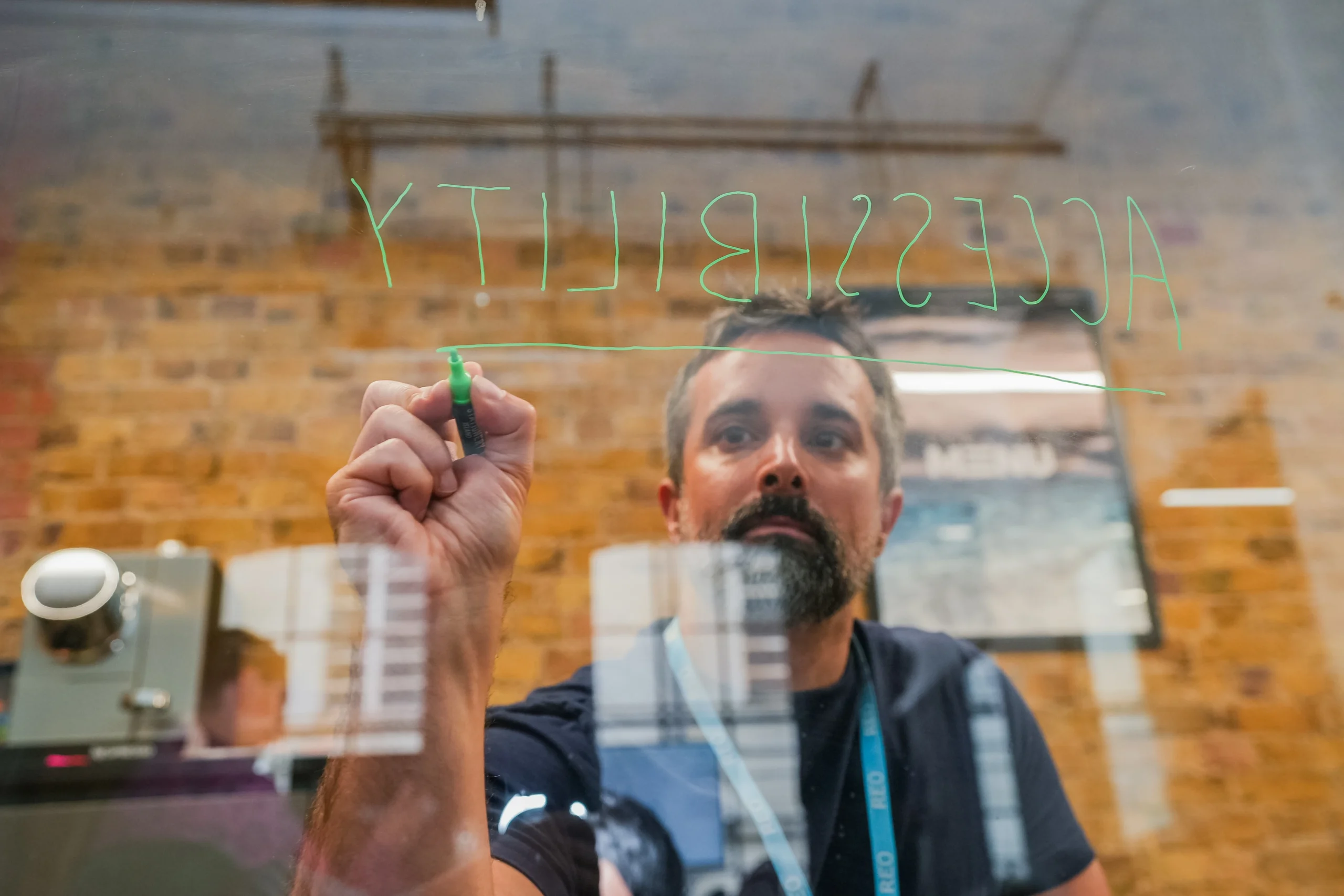 A man writes the word “ACCESSIBILITY” in green marker on a transparent board, viewed from behind the glass.