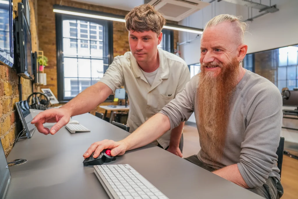 Two men using a desktop computer and adaptive equipment in a workshop setting, one pointing at the screen while the other smiles and interacts with a trackball mouse.