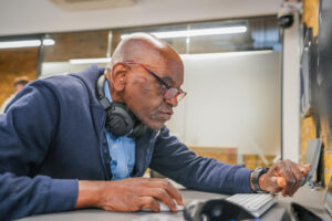 Older man with red glasses and headphones around his neck working intently at a computer in a modern office setting.