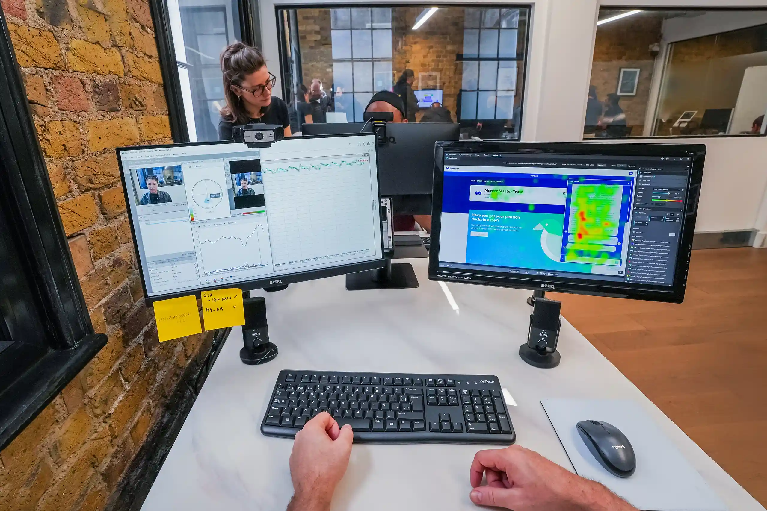 A person sits at a desk with two computer monitors displaying graphs, video feeds, and a heat map while colleagues collaborate in the background through a glass wall.