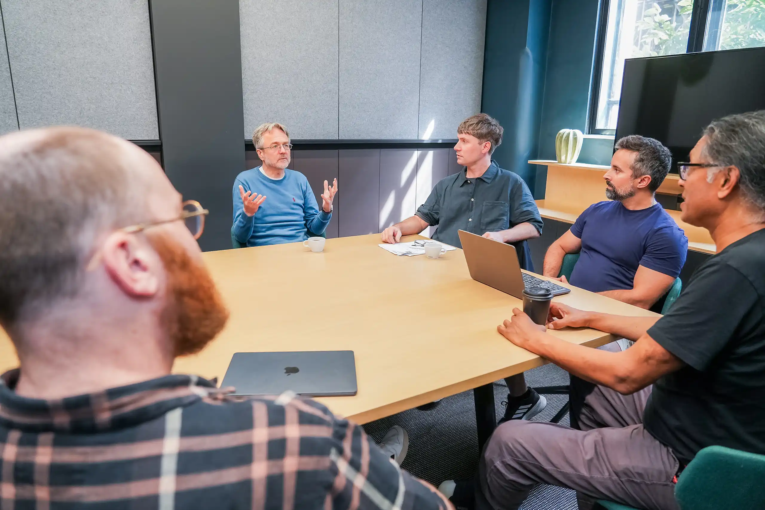 Five colleagues in a meeting room having a discussion around a wooden conference table; one man in a blue sweater is speaking while others listen and take notes on laptops and notepads.